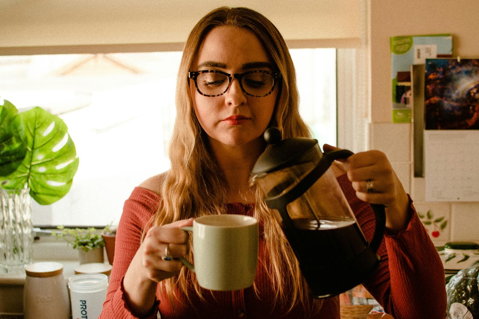A woman pours coffee into a mug using a French press in a cozy kitchen setting.